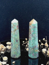 A tall, polished obelisk made of Ruby Fuchsite, standing upright with smooth, transparent facets that catch and reflect light