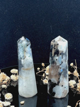 A tall, polished obelisk made of Rainbow Moonstone, standing upright with smooth, transparent facets that catch and reflect light
