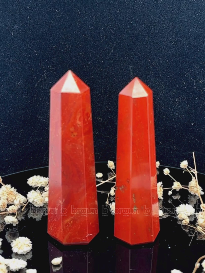 A tall, polished obelisk made of Red Jasper, standing upright with smooth, transparent facets that catch and reflect light