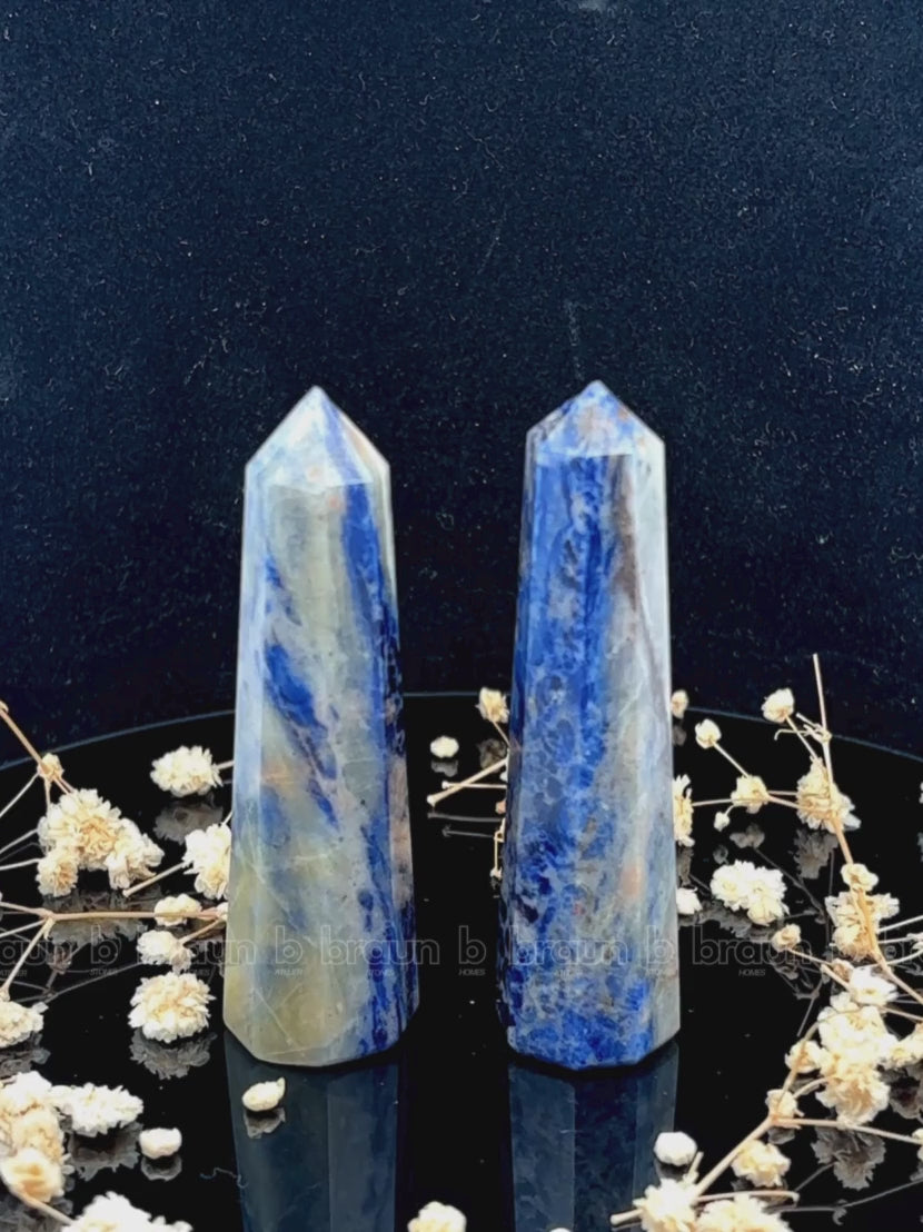 A tall, polished obelisk made of Sodalite, standing upright with smooth, transparent facets that catch and reflect light