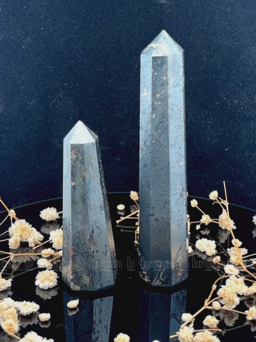 A tall, polished obelisk made of Hematite, standing upright with smooth, transparent facets that catch and reflect light