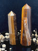 A tall, polished obelisk made of Tiger eye, standing upright with smooth, transparent facets that catch and reflect light