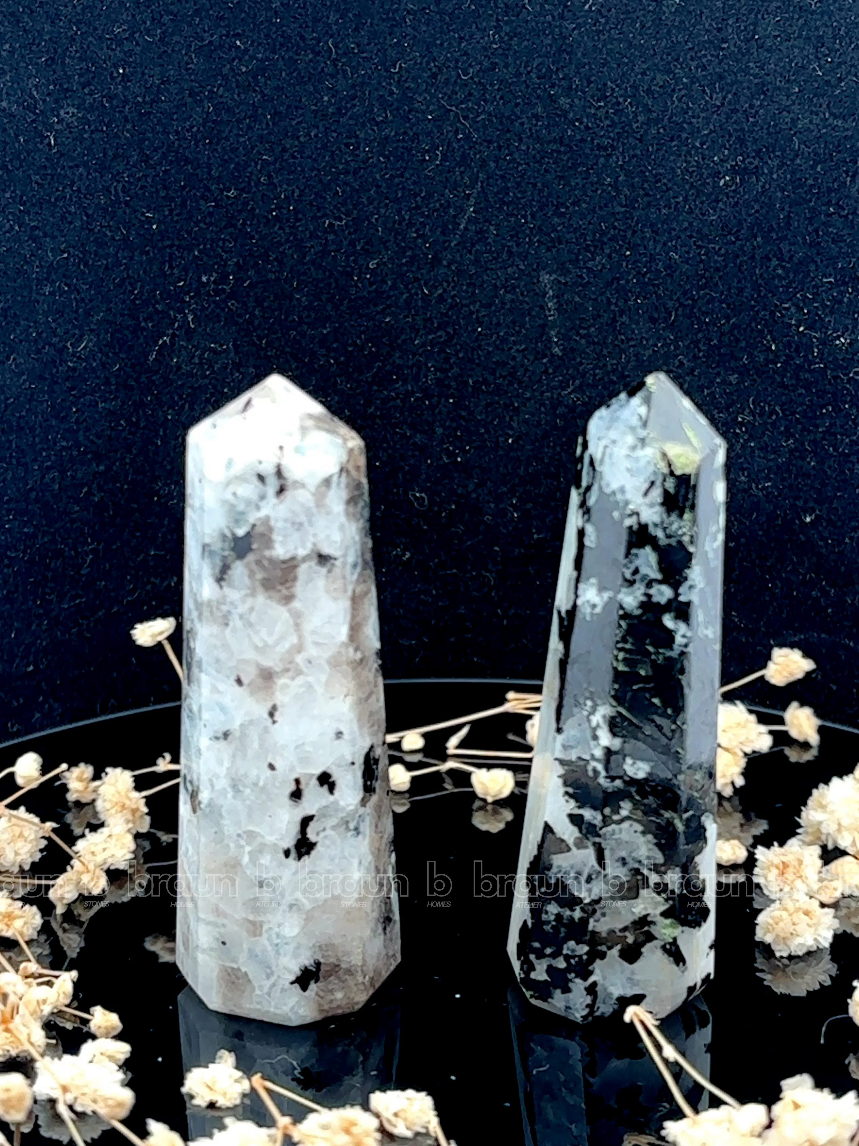 A tall, polished obelisk made of Rainbow Moonstone, standing upright with smooth, transparent facets that catch and reflect light