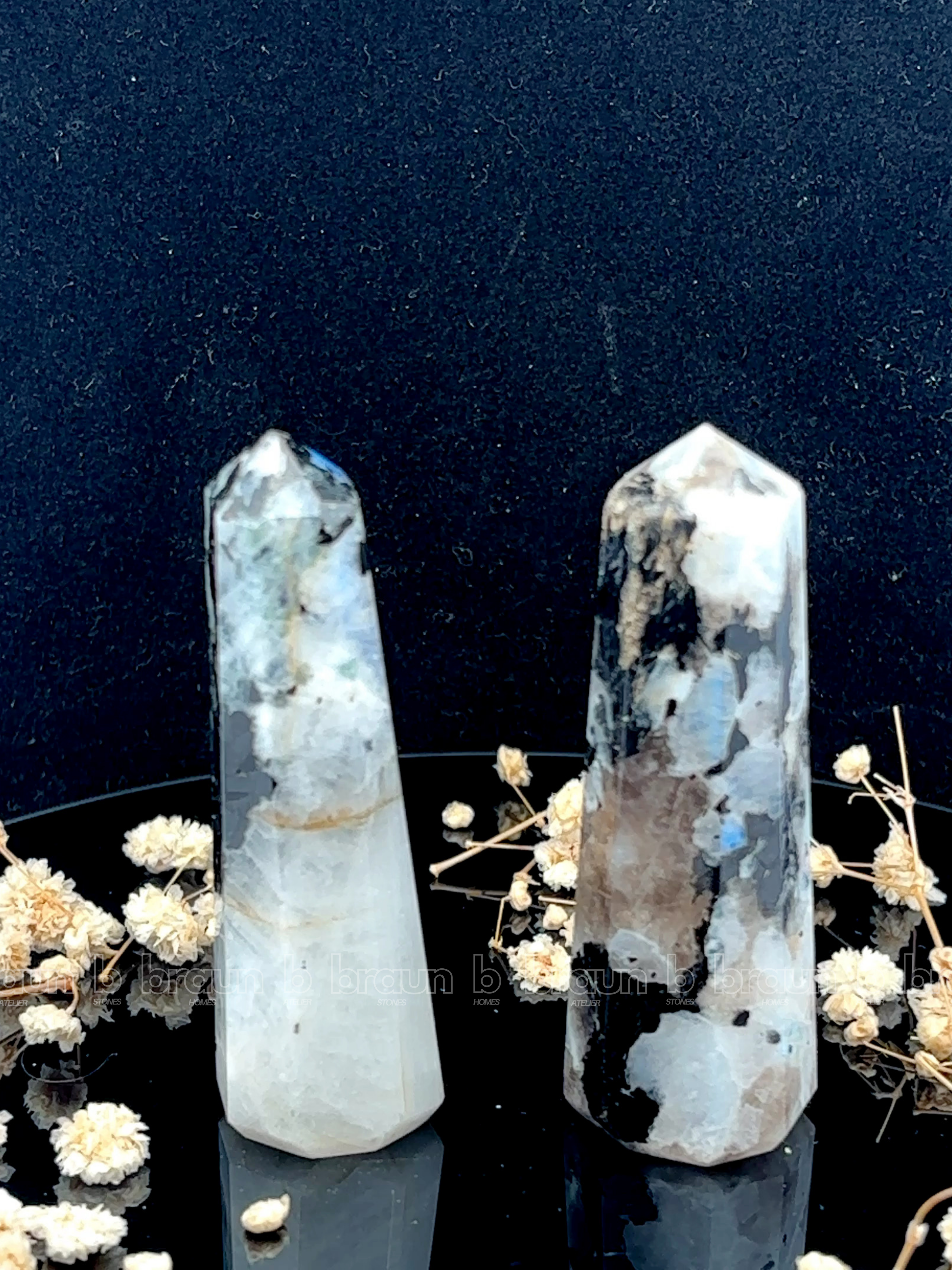 A tall, polished obelisk made of Rainbow Moonstone, standing upright with smooth, transparent facets that catch and reflect light