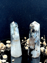 A tall, polished obelisk made of Rainbow Moonstone, standing upright with smooth, transparent facets that catch and reflect light