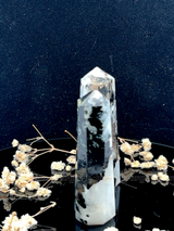 A tall, polished obelisk made of Rainbow Moonstone, standing upright with smooth, transparent facets that catch and reflect light
