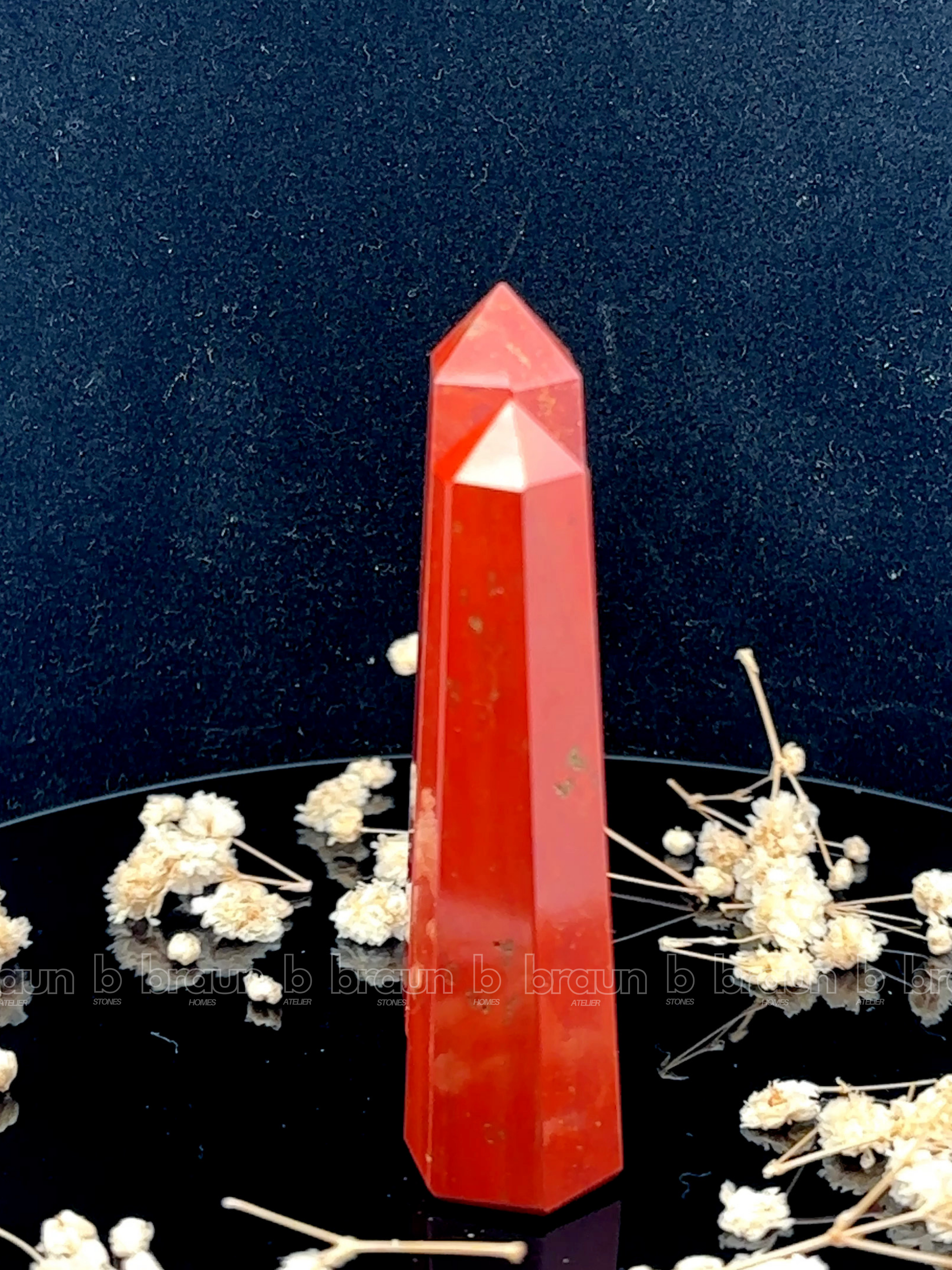 A tall, polished obelisk made of Red Jasper, standing upright with smooth, transparent facets that catch and reflect light