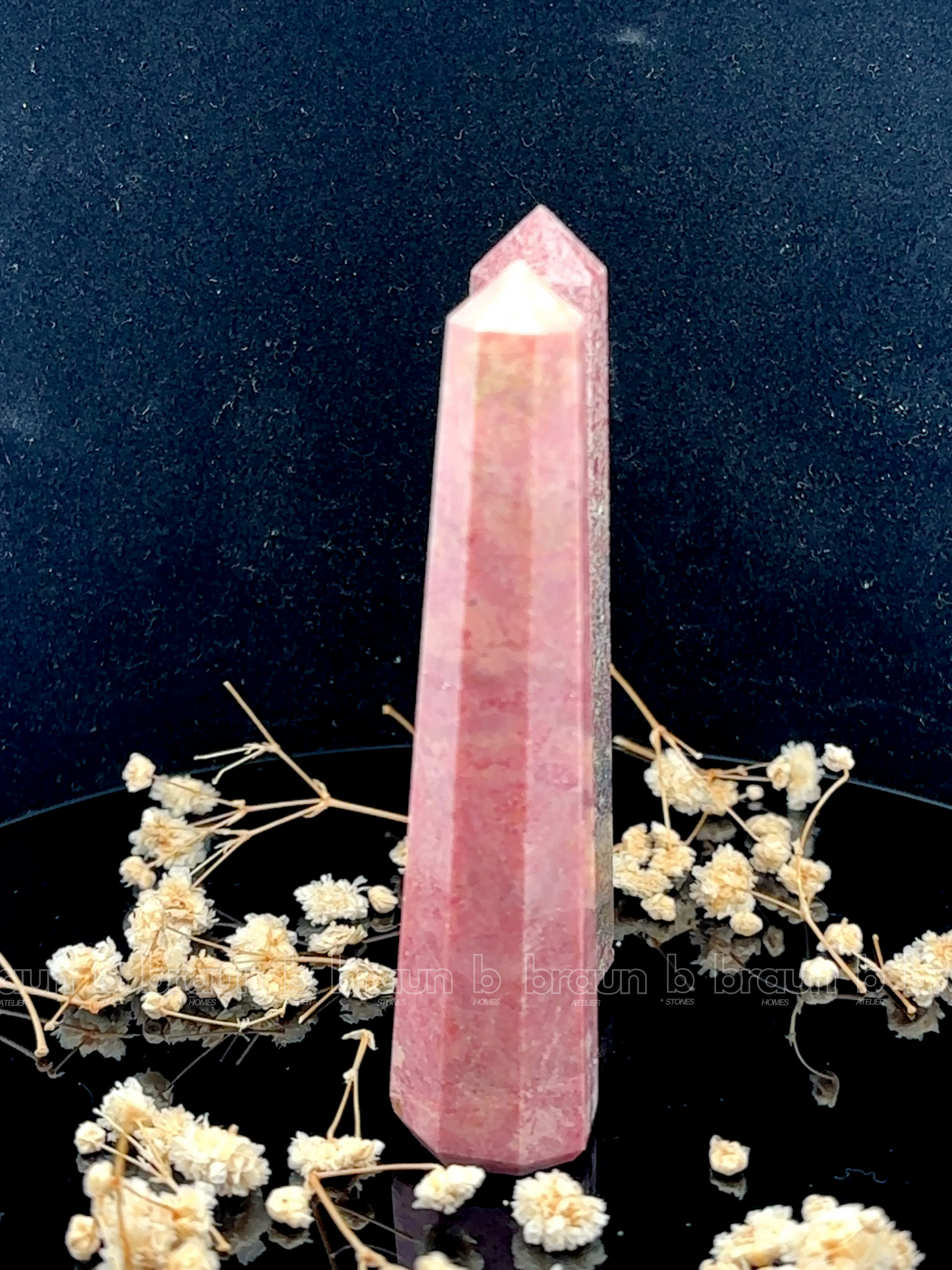 A tall, polished obelisk made of Rhodonite, standing upright with smooth, transparent facets that catch and reflect light