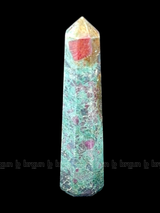 A tall, polished obelisk made of Ruby Fuchsite, standing upright with smooth, transparent facets that catch and reflect light