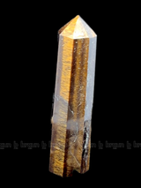 A tall, polished obelisk made of Tiger eye, standing upright with smooth, transparent facets that catch and reflect light