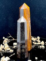 A tall, polished obelisk made of Tiger eye, standing upright with smooth, transparent facets that catch and reflect light