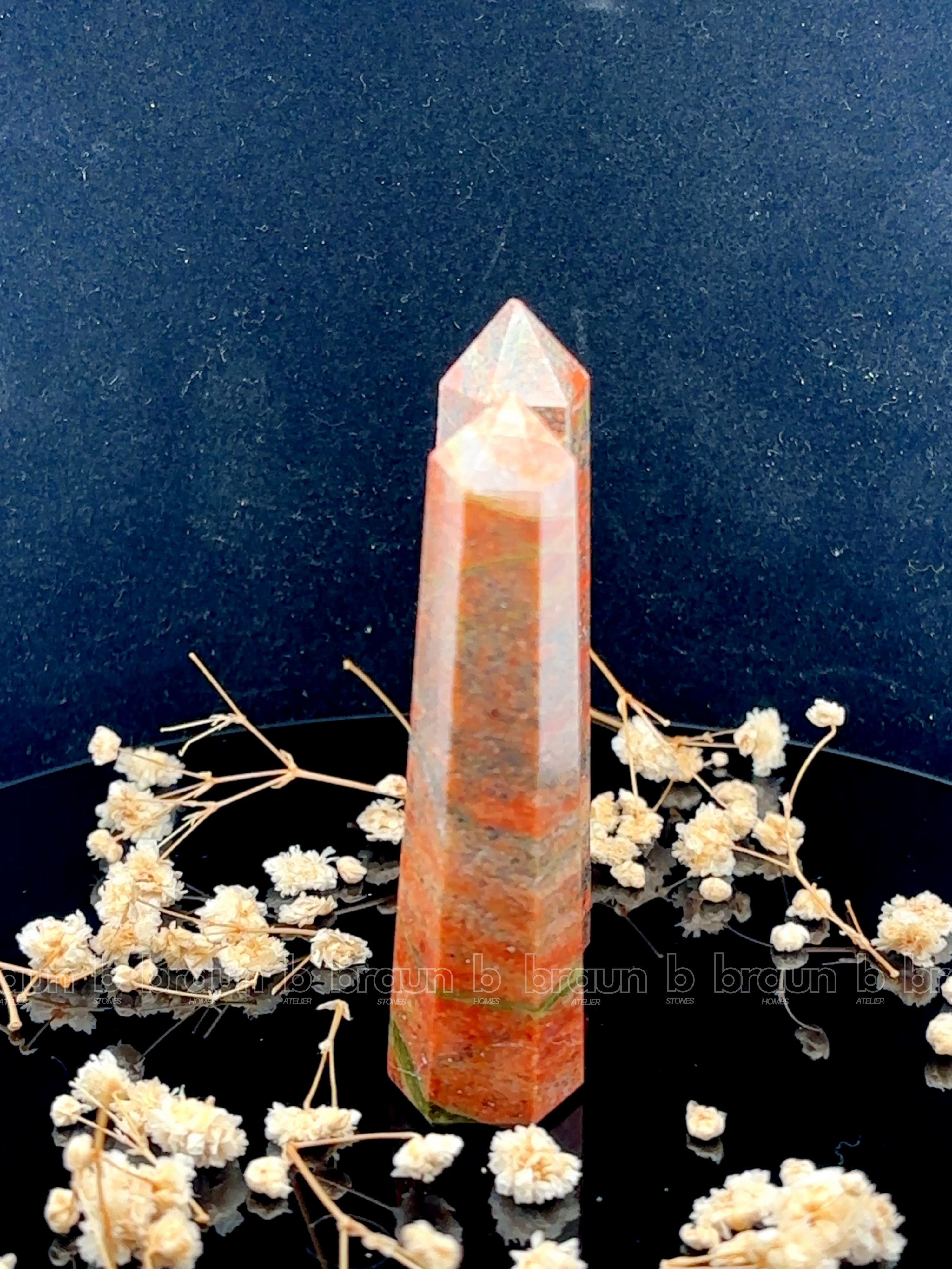A tall, polished obelisk made of Unakite, standing upright with smooth, transparent facets that catch and reflect light
